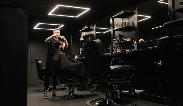 Photo de profil d'un jeune coiffeur coupant les cheveux de son client avec un rasoir électrique et un peigne dans un salon de coiffure.