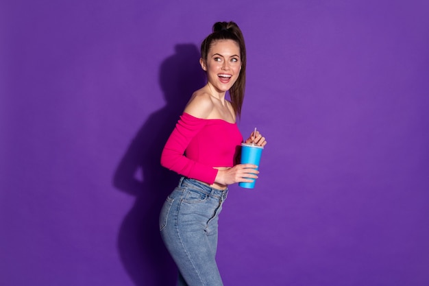 Photo de profil d'une jeune fille folle tenir une tasse de rêve excité porter des jeans en denim rose épaules découvertes haut fond de couleur violet isolé