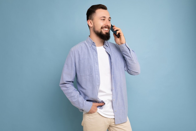 Photo de profil latéral d'un beau jeune homme barbu brun portant une chemise bleue décontractée et un t-shirt blanc en équilibre isolé sur fond bleu avec un espace vide tenant dans la main et communiquant une conversation