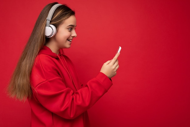 Photo de profil latéral d'une jolie fille brune souriante et positive portant un sweat à capuche rouge isolé sur rouge