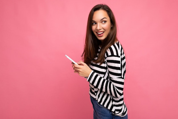 Photo de profil latéral d'une jolie jeune femme brune souriante utilisant un téléphone portable communiquant par SMS portant un pull isolé sur fond de mur regardant sur le côté. Espace de copie