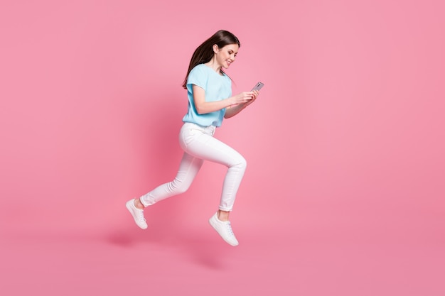 Photo de profil pleine longueur d'une fille sauter utiliser un smartphone courir porter un t-shirt pantalon isolé sur fond de couleur pastel