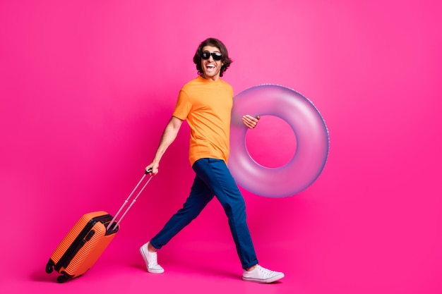 Photo de profil sur toute la longueur de l'homme étape tenir l'anneau de flotteur de bagages porter un t-shirt orange jeans lunettes de soleil chaussures isolé fond de couleur rose