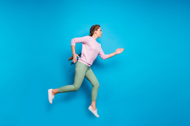 Photo de profil sur toute la longueur de la jolie dame de longues tresses sautant la compétition sportive de haut niveau jusqu'à la ligne d'arrivée porter des pantalons verts pull rose décontracté fond de couleur bleu isolé