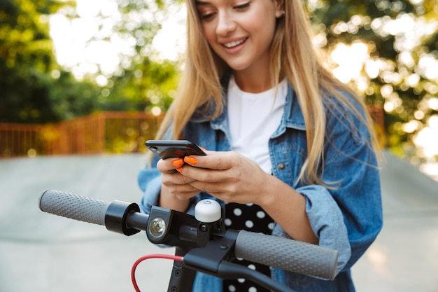 Photo recadrée d'une adolescente heureuse souriante positive dans un parc marchant sur un scooter à l'aide d'un téléphone portable.