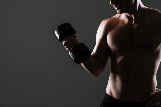 Photo recadrée d'un athlète séduisant debout avec un haltère dans une salle de sport sur un mur gris.