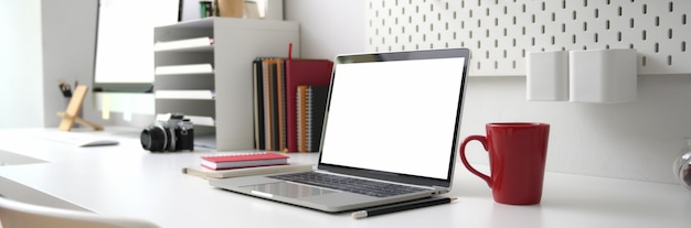 Photo recadrée de bureau avec ordinateur portable à écran blanc, fournitures de bureau et tasse à café