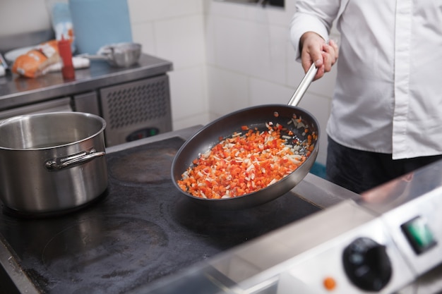 Photo recadrée d'un chef professionnel faisant frire des légumes hachés sur la poêle dans la cuisine du restaurant