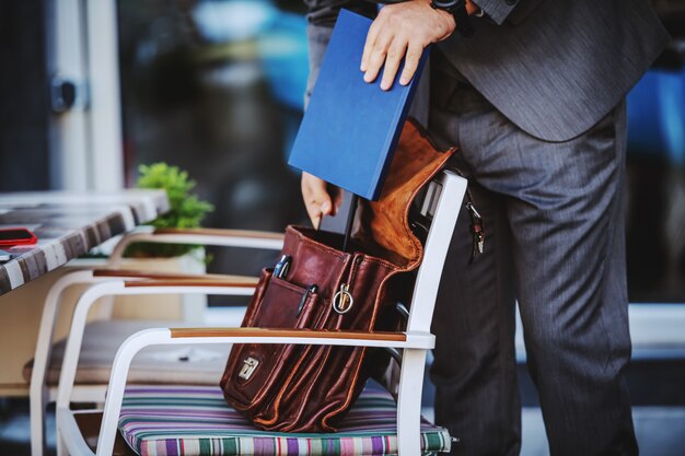 Photo recadrée de l'élégant homme d'affaires en costume sortant l'ordre du jour de son sac en cuir. Extérieur du café.