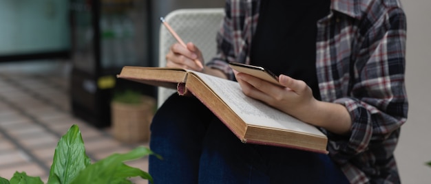 Photo recadrée d'une étudiante à l'aide de smartphone pour rechercher des informations tout en lisant un livre dans le jardin