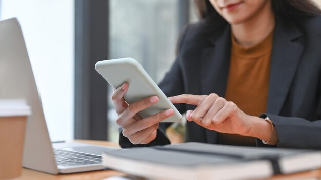 Photo recadrée d'une femme d'affaires heureuse utilisant un téléphone intelligent alors qu'elle était assise dans un bureau moderne.