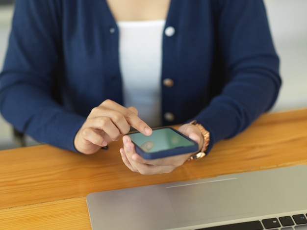 Photo recadrée de femme à l'aide de smartphone tout en travaillant avec un ordinateur portable sur un bureau en bois