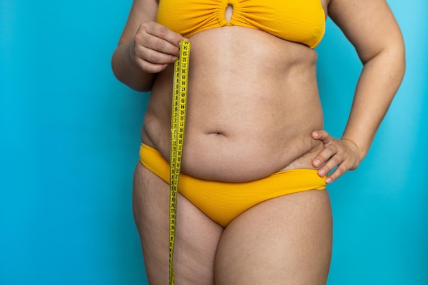 Photo recadrée d'une femme dodue debout dans un maillot de bain jaune montrant un excès de ventre nu tenant un ruban à mesurer