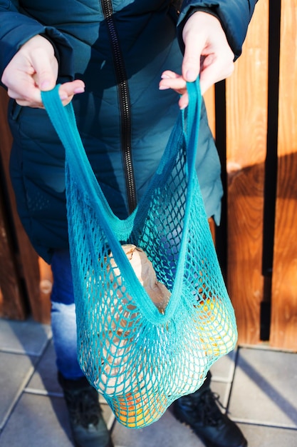 Photo recadrée d'une femme méconnaissable tenant un sac en filet bleu tricoté Photo extérieure sac tricoté plein de fruits et légumes dans les mains d'une femme Planches de bois sur fond Espace de copie