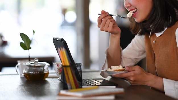 Photo recadrée d'une femme travaillant avec une tablette informatique et mangeant un dessert au café.