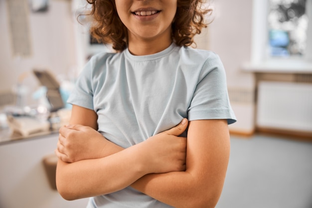 Photo recadrée d'un garçon souriant bouclé en chemise grise debout, les bras croisés dans une clinique dentaire