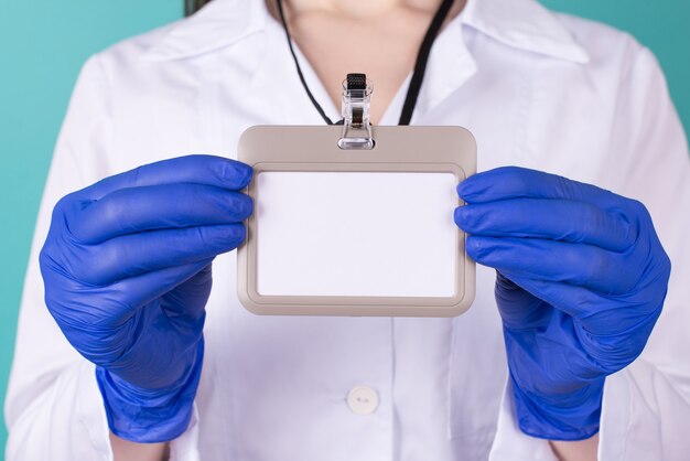 Photo photo recadrée en gros plan d'une femme médecin portant des gants d'uniforme en blouse blanche tenant une étiquette de nom de maquette vierge devant isolée sur fond bleu turquoise