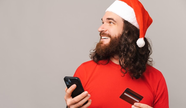 Photo recadrée d'un homme barbu joyeux portant un bonnet rouge de santa tenant un smarphone et une carte de crédit et regardant ailleurs