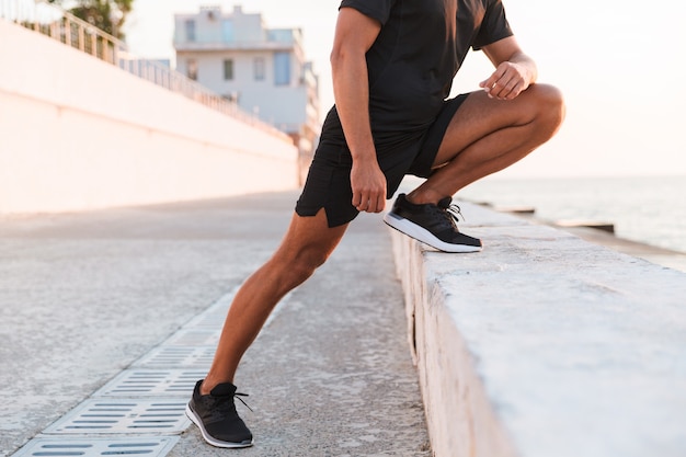 Photo recadrée d'un homme fort de 30 ans en short et t-shirt faisant du sport près de la mer et s'étirant les jambes au lever du soleil