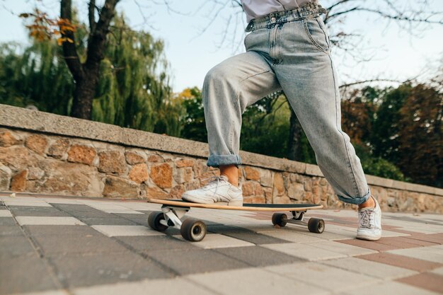 Photo recadrée de jambes de skateur féminin en jeans baggy chevauchant sa longue planche dans une rue de la ville