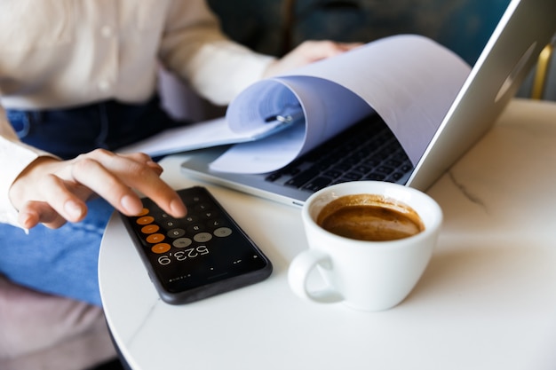 Photo recadrée de jeune femme assise dans un café à l'intérieur de travailler avec un ordinateur portable contenant des documents à l'aide d'un téléphone mobile.