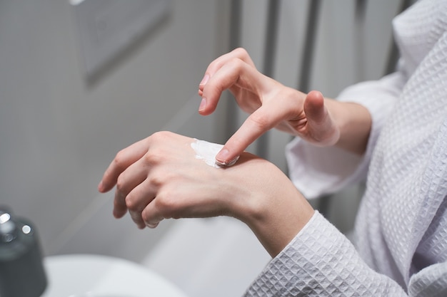 Photo recadrée d'une jeune femme de race blanche dans un peignoir appliquant une crème hydratante pour les mains