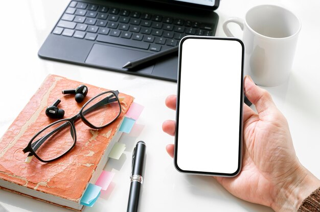 Photo recadrée d'une main d'homme tenant un smartphone à écran blanc avec un bureau en arrière-plan.