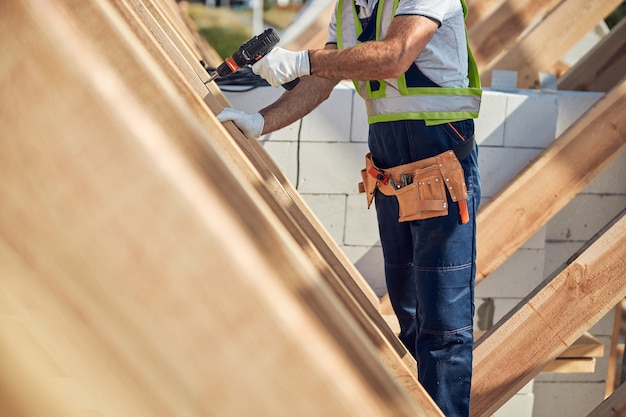 Photo recadrée d'un ouvrier de chantier portant une ceinture à outils pratique et perçant le cadre de toit en bois