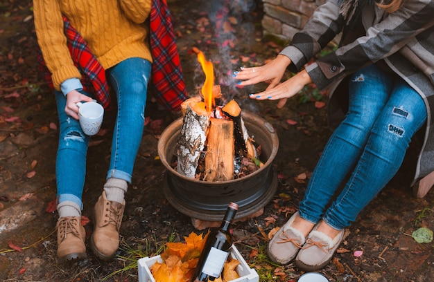 Photo recadrée de trois femmes meilleures amies assises autour d'un feu de joie dans des vêtements décontractés s'échauffant et communiquant, tenant des tasses avec des boissons chaudes, heureuses d'être ensemble dehors par une froide soirée d'automne