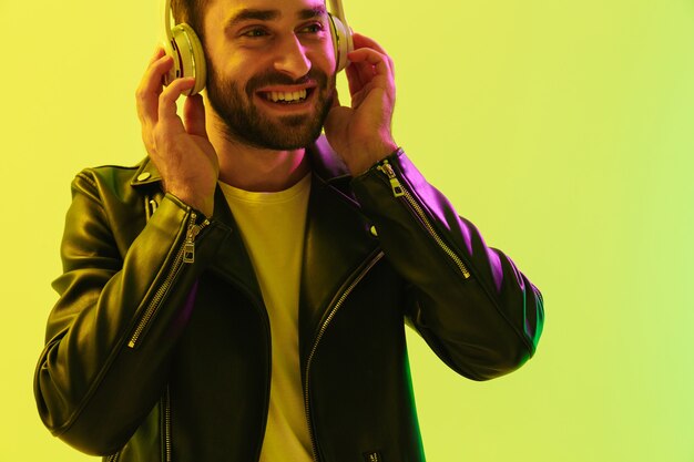 Photo de sourire heureux beau jeune homme élégant posant isolé sur un mur vert clair avec des néons à led vêtus d'une veste en cuir écoutant de la musique avec des écouteurs.