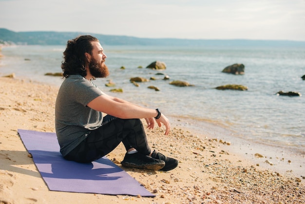 Photo d'un sportif barbu assis sur un tapis près du bord de mer pendant la matinée