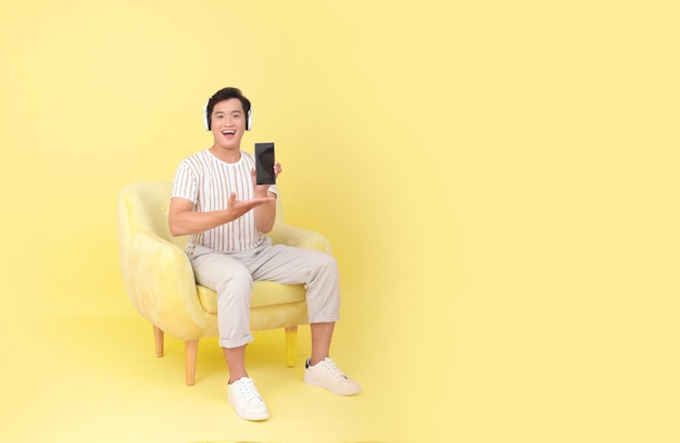 Photo de stock de corps pleine longueur d'un beau jeune homme asiatique assis sur une chaise de canapé jaune isolé sur fond jaune montrant l'écran du téléphone intelligent Concept pour les gadgets de haute technologie de l'utilisateur de l'application ou la promotion Internet