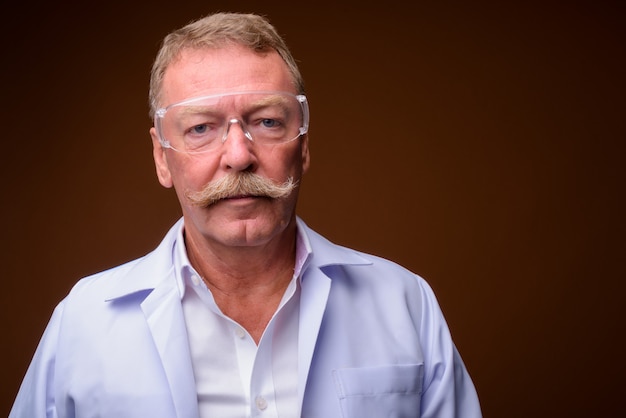 Photo de Studio de bel homme senior médecin avec moustache portant des lunettes de protection contre brown