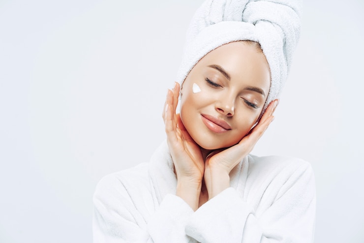 Photo photo de studio de belle femme détendue avec les yeux fermés incline la tête, touche la peau doucement, applique la crème pour le visage, porte une serviette de bain sur la tête après avoir pris des procédures de spa, bénéficie de soins du visage, soins du corps