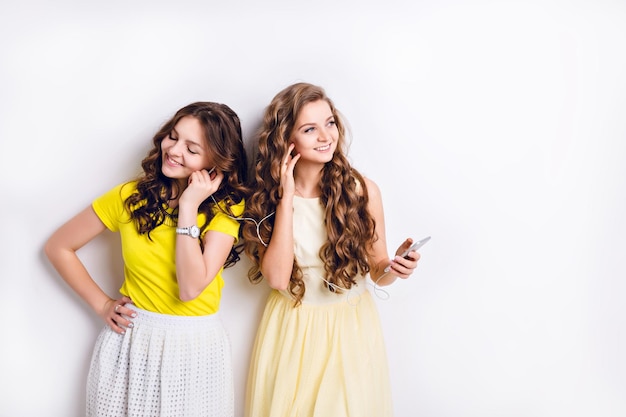 Photo de studio de deux filles souriantes debout écoutant de la musique sur un smartphone et s'amusant. La brune porte une jupe blanche et un t-shirt jaune, et la fille blonde porte une robe jaune.