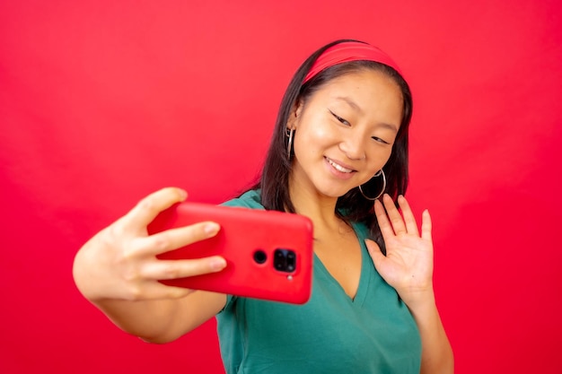 Photo de studio avec un fond rouge d'une femme chinoise agitant tout en prenant un selfie