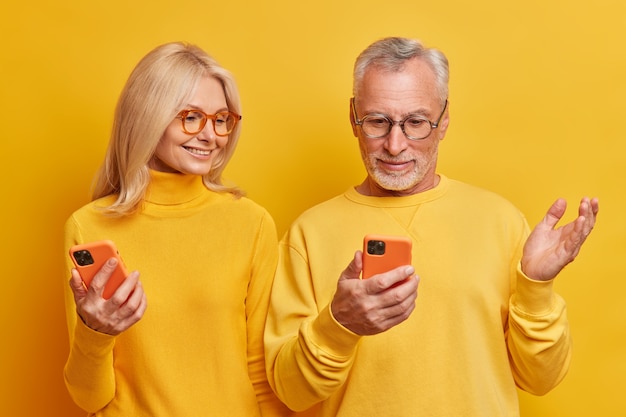 Photo de Studio d'un homme âgé semble perplexe devant l'affichage du smartphone a un problème soulève la paume de sa femme essaie de l'aider à se tenir étroitement l'un à l'autre isolé sur un mur jaune
