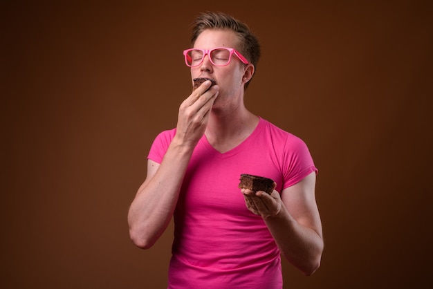 Photo de Studio de jeune bel homme portant une chemise rose avec des lunettes roses assorties tout en ayant un gâteau au chocolat sur fond marron