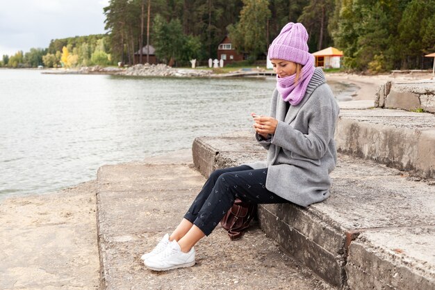 Photo de style de vie atmosphérique en plein air d'une belle jeune femme brune en bonnet à tricoter, dans un manteau gris et un pantalon noir, assise sur la jetée à côté de la mer par une journée ensoleillée d'automne