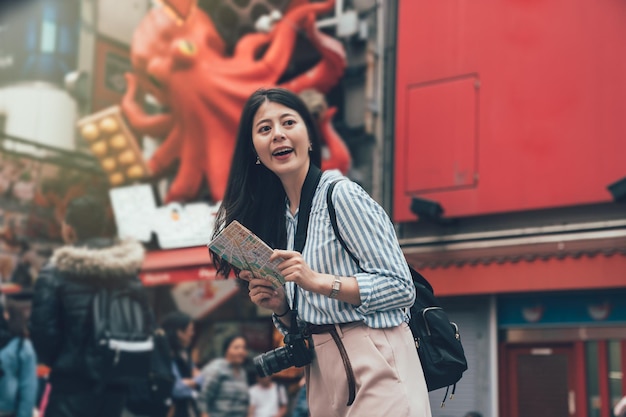 photo de style vintage d'un photographe de sac à dos gril souriant debout devant le magasin de vendeurs de nourriture takoyaki avec une énorme publicité d'animaux de poulpe accrochée au mur en arrière-plan. Dame gaie avec carte