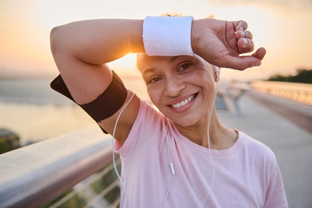 Photo de tête d'une belle athlète hispanique essuyant la sueur de son front avec un bracelet en éponge, sourit avec un sourire joyeux à pleines dents se reposant après le jogging du matin sur un tapis roulant de pont de la ville au lever du soleil
