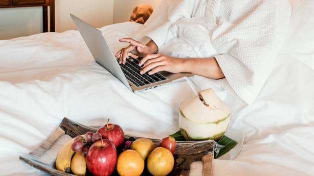 Photo de touristes utilisant un ordinateur portable et mangeant des fruits sur le lit, concept d'aliments santé.