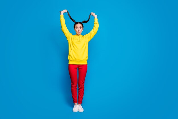 Photo de tout le corps d'une dame assez gentille et drôle tenir les bras à deux queues bonne humeur étonnée croissance rapide cheveux porter un pull à capuche jaune décontracté pantalon rouge baskets isolé fond de couleur bleu vif