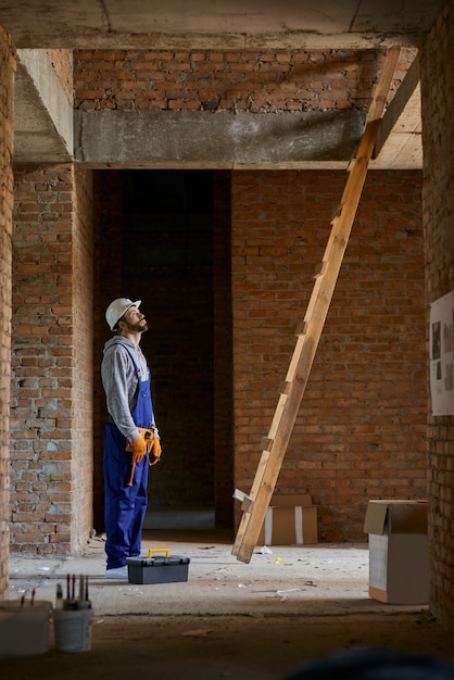 Photo de toute la longueur d'un jeune ouvrier en salopette bleue et casque de protection en levant les yeux tout en montant l'échelle, travaillant sur la construction d'une maison