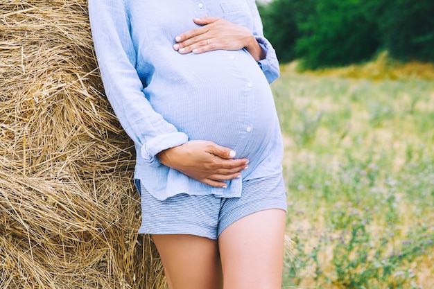 Photo de ventre de femme enceinte dans la nature Femme enceinte tient les mains sur le ventre sur fond naturel de champ de blé avec des meules de foin au jour d'été Concept de grossesse maternité Gros plan à l'extérieur