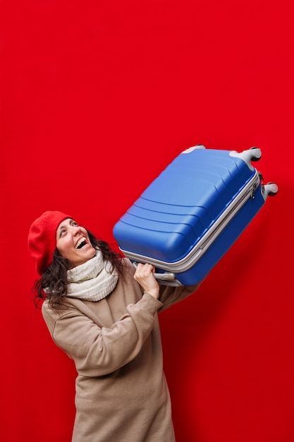 Photo verticale d'une belle femme élégante à la recherche d'une valise bleue pour voyager avec un mur de voyage rouge isolé.