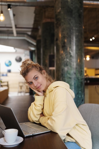 Photo verticale d'une belle fille assise dans un café avec un ordinateur portable, boire du café Étudiante étudiant à distance s'inscrire à des cours en ligne et travaillant sur un projet depuis un café Femme souriante travaillant au coworking