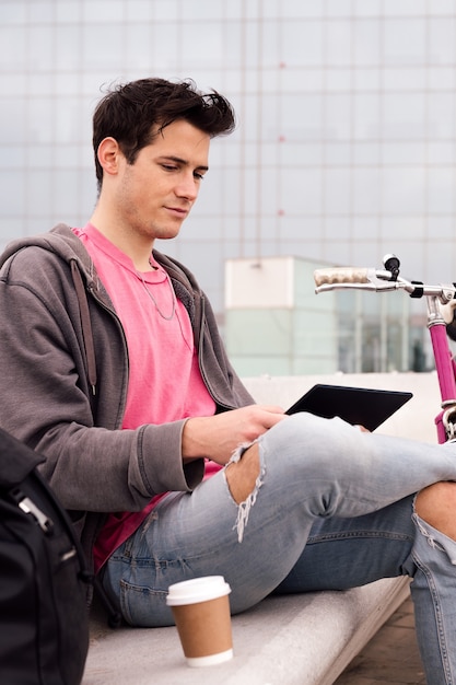 Photo verticale d'un étudiant assis à l'extérieur à l'aide d'une tablette à côté de son concept de technologie à vélo