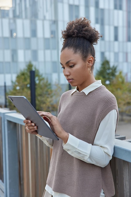 Photo la photo verticale d'un étudiant intelligent porte des vêtements soignés utilise une tablette numérique vérifie les flux dans les réseaux sociaux parcourt le site web recherche des informations pour les poses de projet sur le pont sur fond urbain