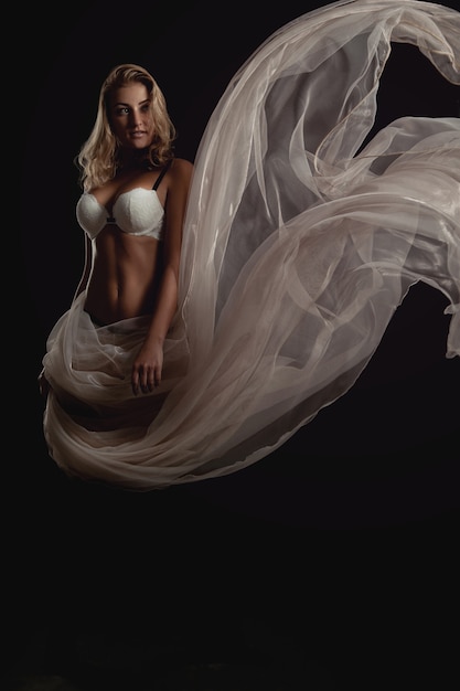 Photo verticale d'une femme blonde sexuelle en soutien-gorge blanc et tissu volant en studio sur fond noir